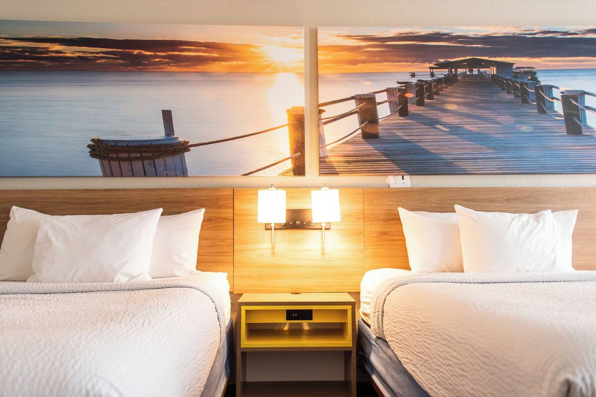 A hotel room with two beds, a wooden headboard, and a painting of a pier at sunset.