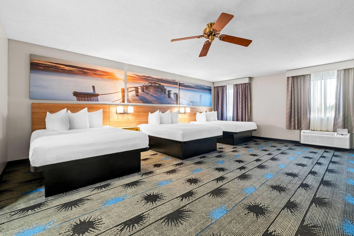 A hotel room with a large bed, two smaller beds, a ceiling fan, a painting of a sunset, and a view of the sea.
