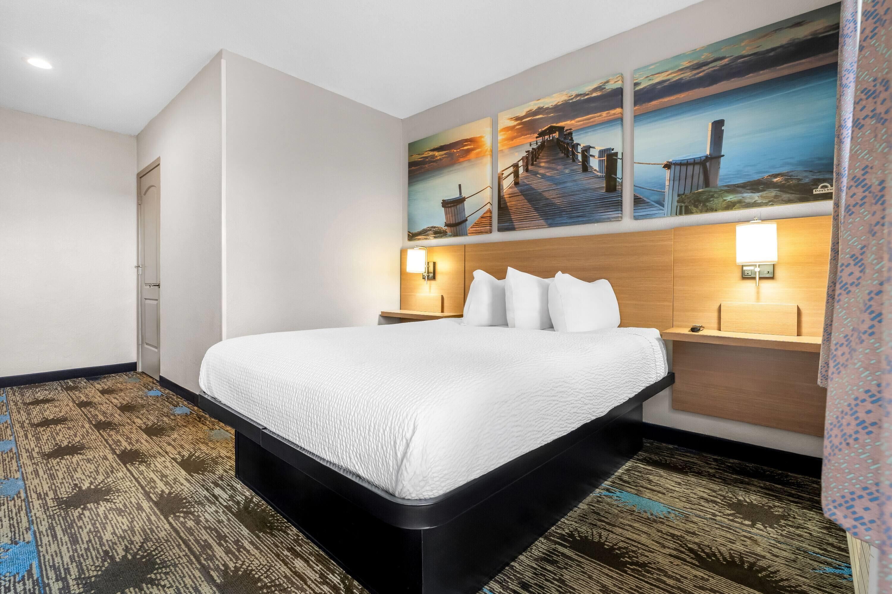 Habitación de hotel con una cama, dos mesitas de noche con lámparas, un cabecero de madera y una pared con tres cuadros.
