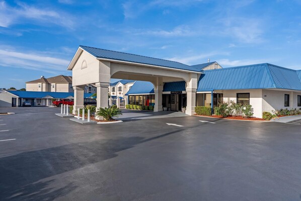 A hotel with a covered entrance, a parking lot, and a blue-roofed building in the background.