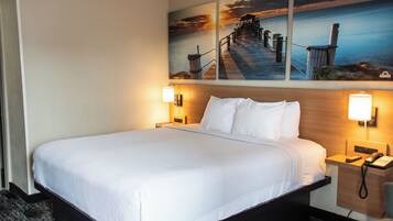 A hotel room with a large bed, two bedside tables, a nightstand, and a painting of a pier and sunset.