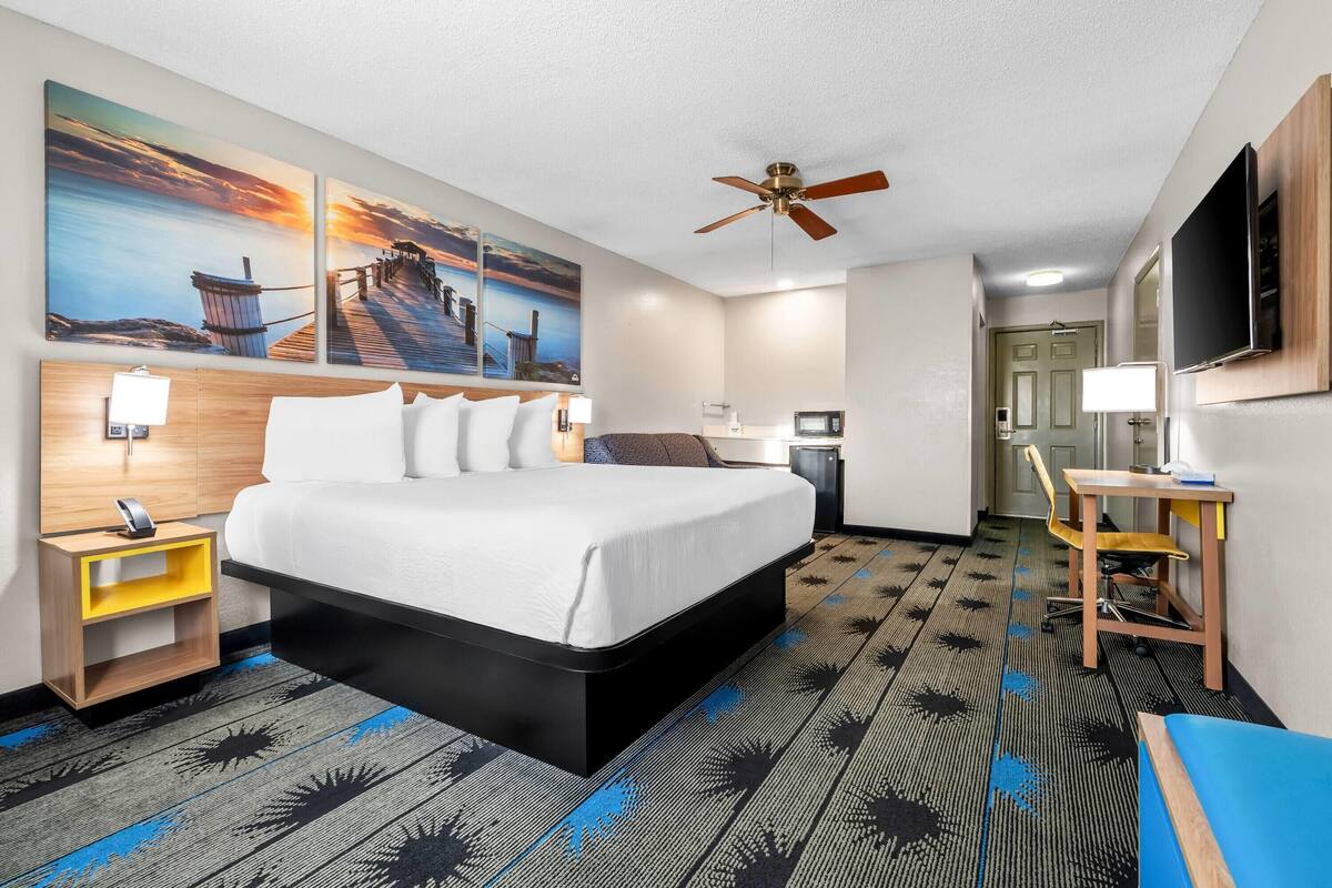 A hotel room with a bed, a desk, a chair, a ceiling fan, and artwork on the wall.