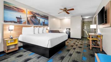 A hotel room with a bed, a desk, a chair, a ceiling fan, and artwork on the wall.