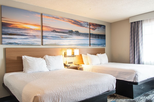 Une chambre d’hôtel avec deux lits, une tête de lit en bois, une table de chevet avec une lampe et un grand tableau représentant un coucher de soleil, placé au-dessus des lits.