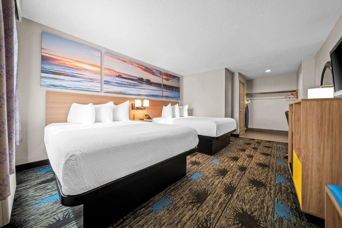 A hotel room with two beds, a large painting of a sunset, a wooden headboard, and a patterned carpet.