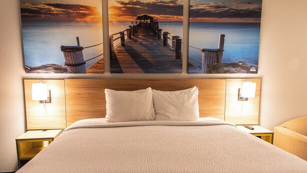 Un cuarto de hotel con una cama grande, dos lámparas de noche y una vista del atardecer en la pared.