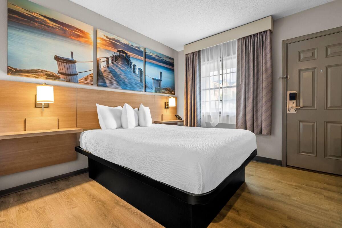 A hotel room with a large bed, wooden headboard, two wall-mounted lamps, a door with a lock, and a large window with curtains.