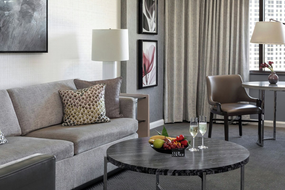 A modern hotel room with a grey sofa, a round coffee table with fruit and glasses, a chair, and a lamp.