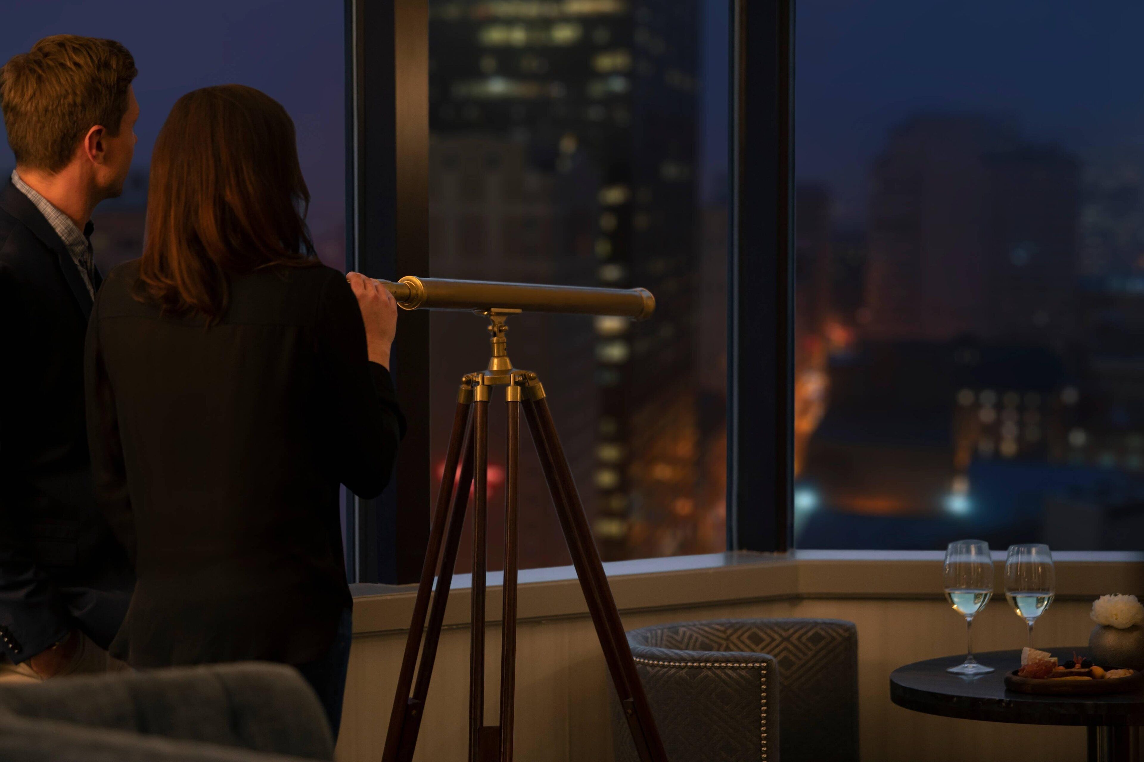 Un couple observe la vue sur la ville depuis une fenêtre. On aperçoit un télescope posé sur un trépied, ainsi qu’une table sur laquelle se trouvent deux verres de vin et un petit vase.
