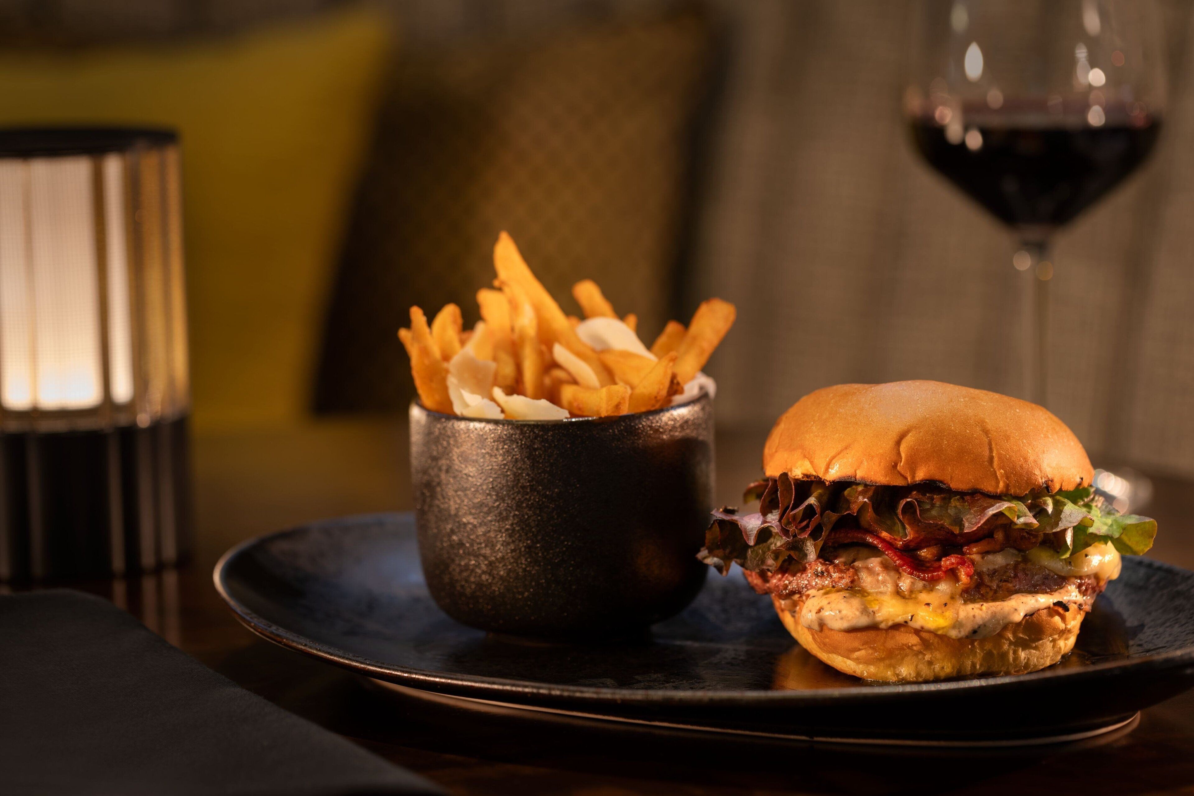A burger and fries on a plate with a glass of red wine in the background.