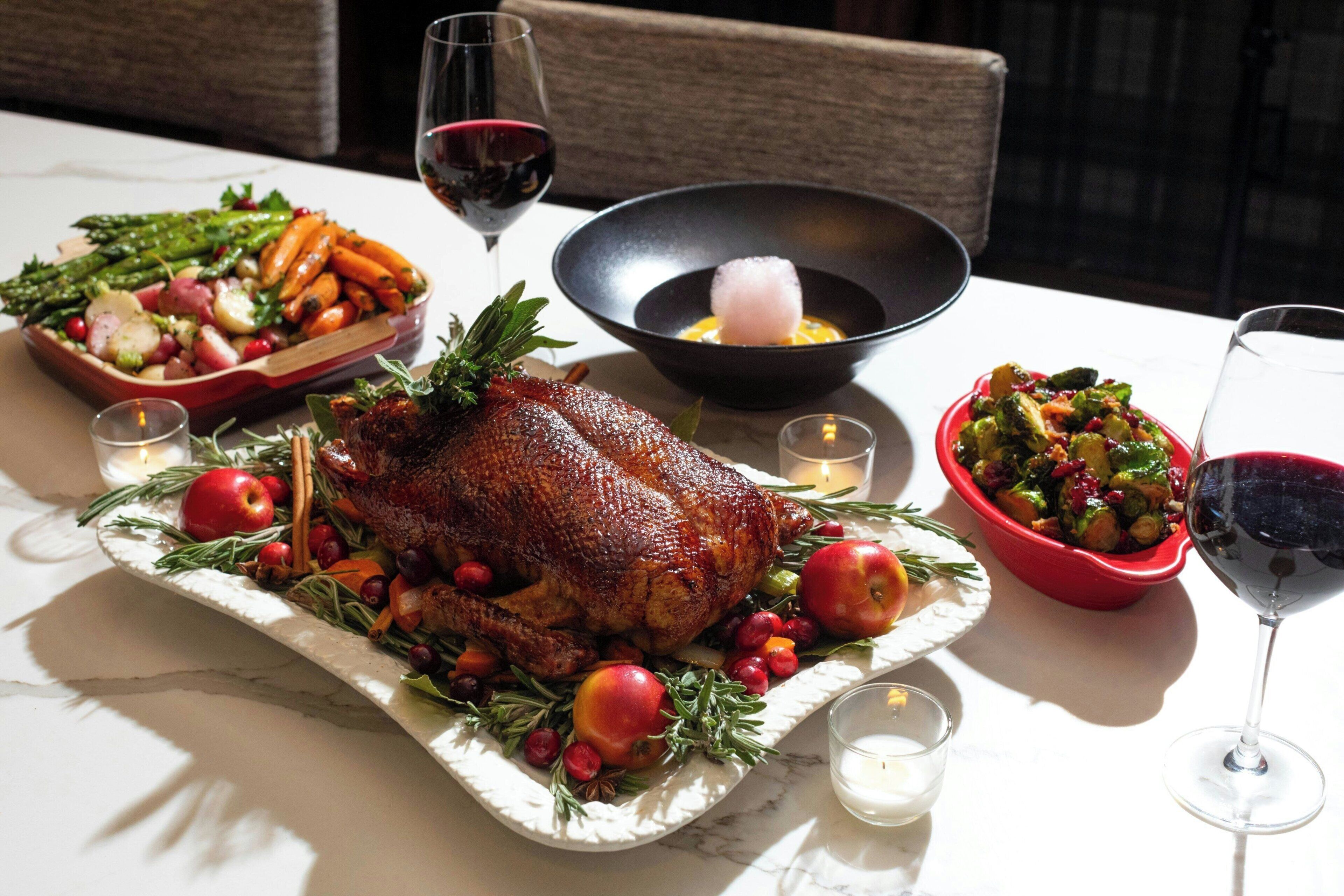 A roasted turkey surrounded by vegetables and fruits on a marble table with candles and wine glasses.