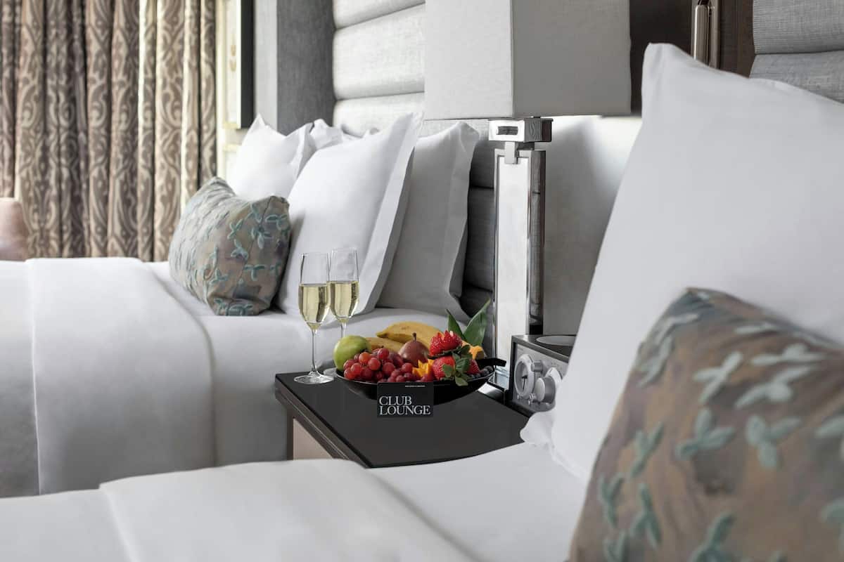 A hotel room with a bed, a bedside table with a fruit bowl, two glasses of wine, and a sign reading "CLUB LOUNGE".