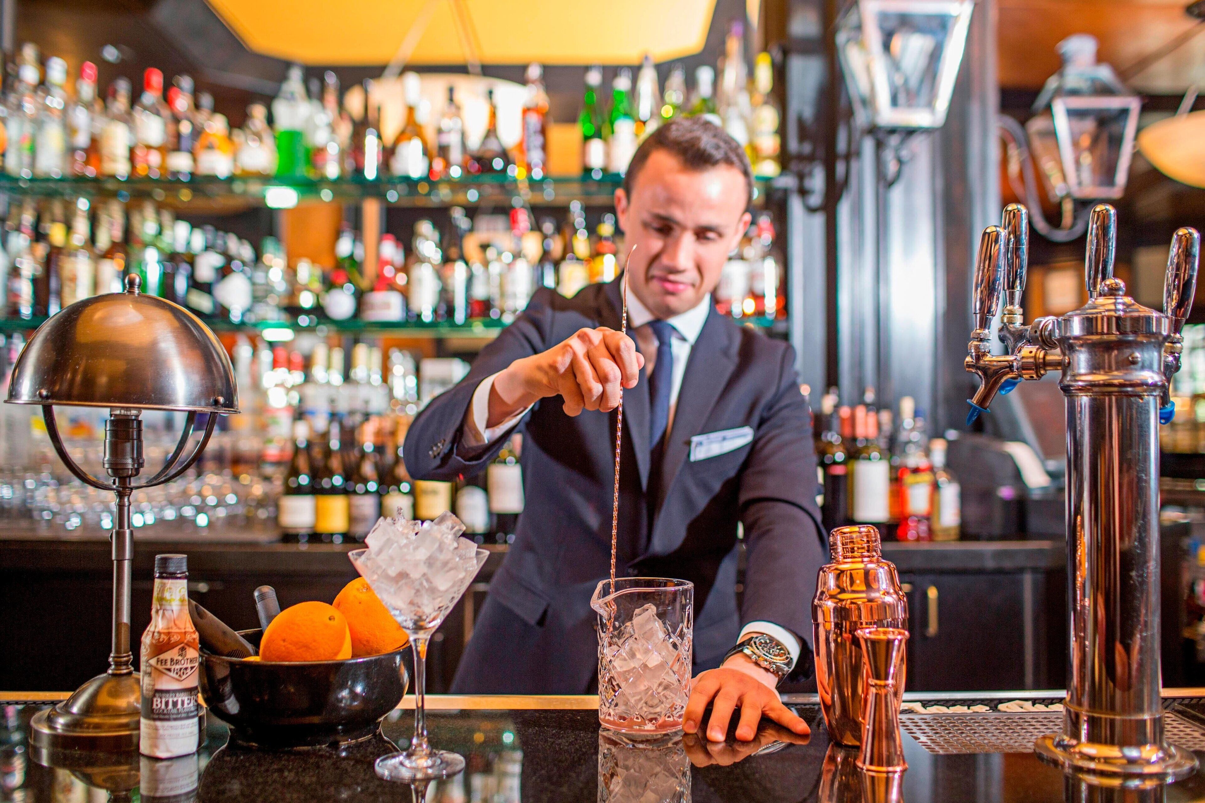 Un barman verse une boisson dans un verre, avec une variété de bouteilles et un narguilé en arrière-plan.