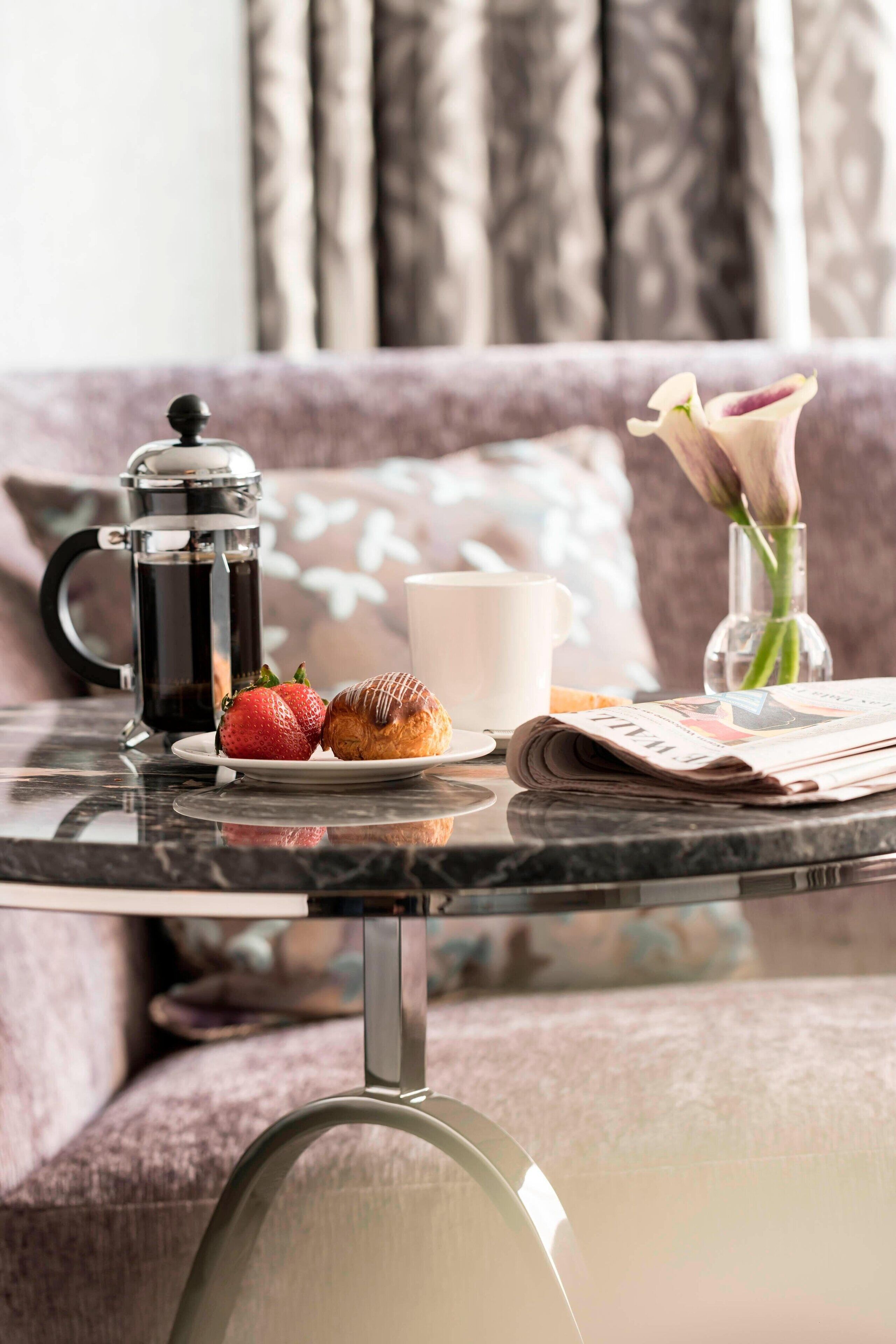 Une table en verre est dressée pour le petit-déjeuner : on y trouve une cafetière, une assiette avec un croissant et des fraises, une tasse et un journal.