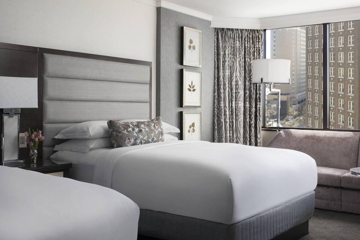 A hotel room with a large bed, a sofa, a nightstand with a lamp, and a view of buildings through the window.