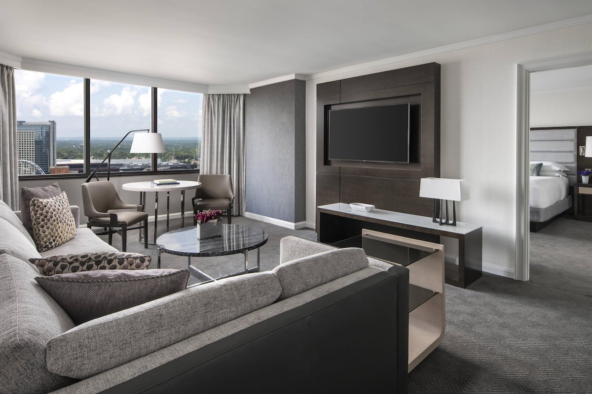 A modern hotel room with a sofa, armchair, coffee table, TV, and a bed.