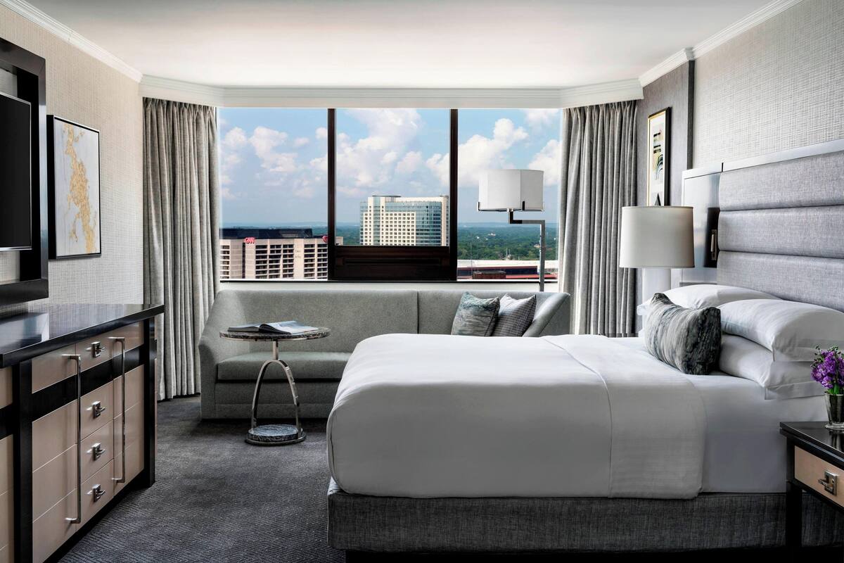 A modern hotel room with a large bed, a sofa, a TV, and a view of the cityscape.