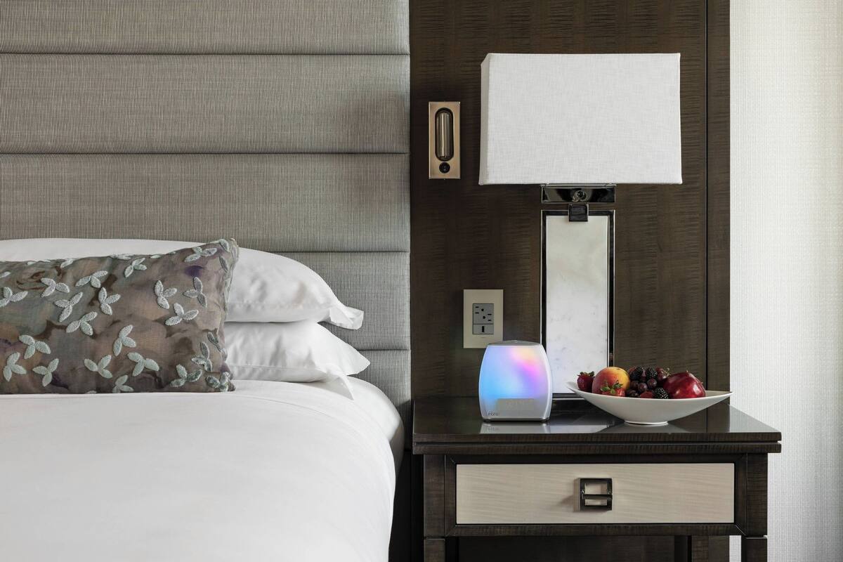 A neatly made bed with a patterned pillow, a bedside table with a bowl of fruit, a lamp, and a wall-mounted light switch.