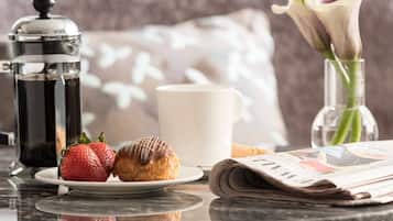 Un desayuno con una cafetera, un plato con una fresa y un pastel, una taza, un florero con una flor y un periódico.