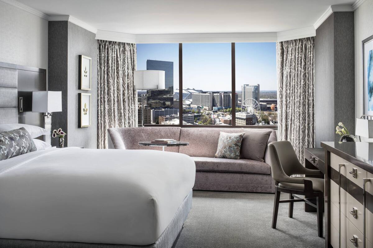 A hotel room with a large bed, a sofa, a desk, and a view of the cityscape through large windows.