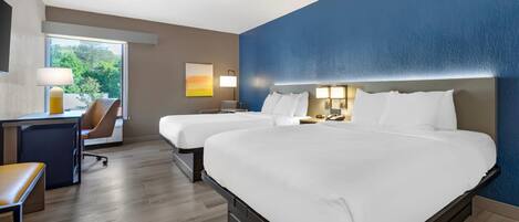 A hotel room with two beds, a desk, a chair, and a window with a view of trees.