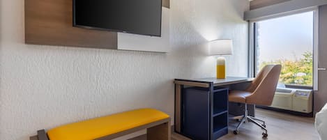 Habitación de hotel con televisión de pantalla plana en la pared, un escritorio con silla, una banca y un ventanal con vistas.