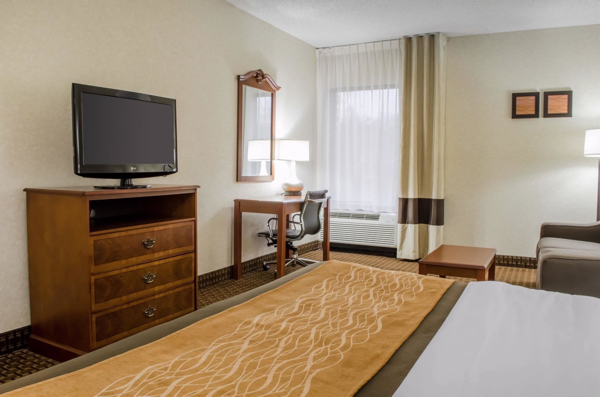 Ein Hotelzimmer mit einem Bett, einem Fernseher auf einer Holzkommode, einem Schreibtisch mit Stuhl, einer Lampe und einem Fenster mit Vorhängen.