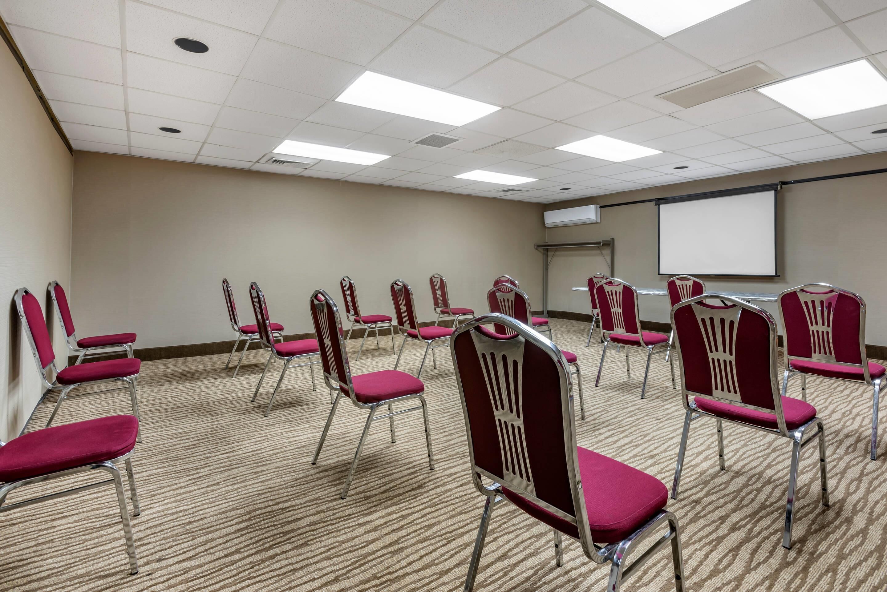 A conference room with rows of red chairs, a carpeted floor, a projector screen, and a whiteboard.