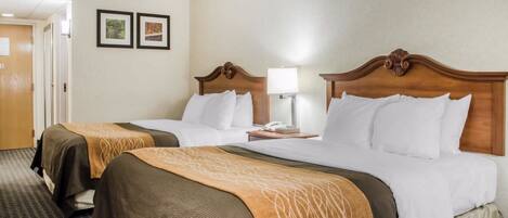 A hotel room with two beds, each with a wooden headboard, white bedding, and a brown decorative throw. There is a bedside table with a lamp and a phone. The room has a carpeted floor and a door leading to another room.