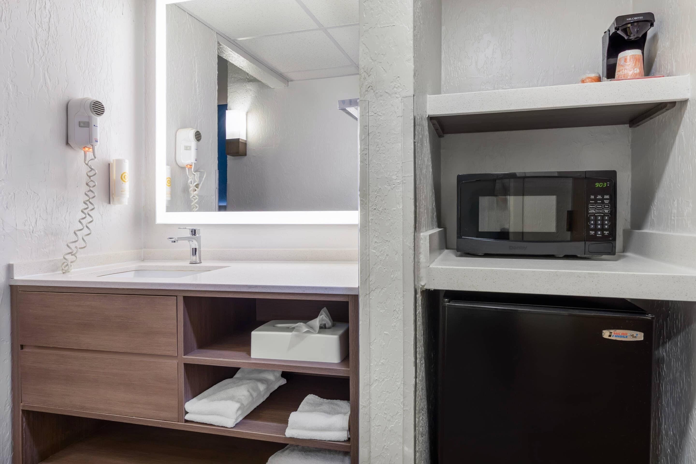 Un baño con lavamanos, secador de pelo, espejo, microondas y refrigerador.