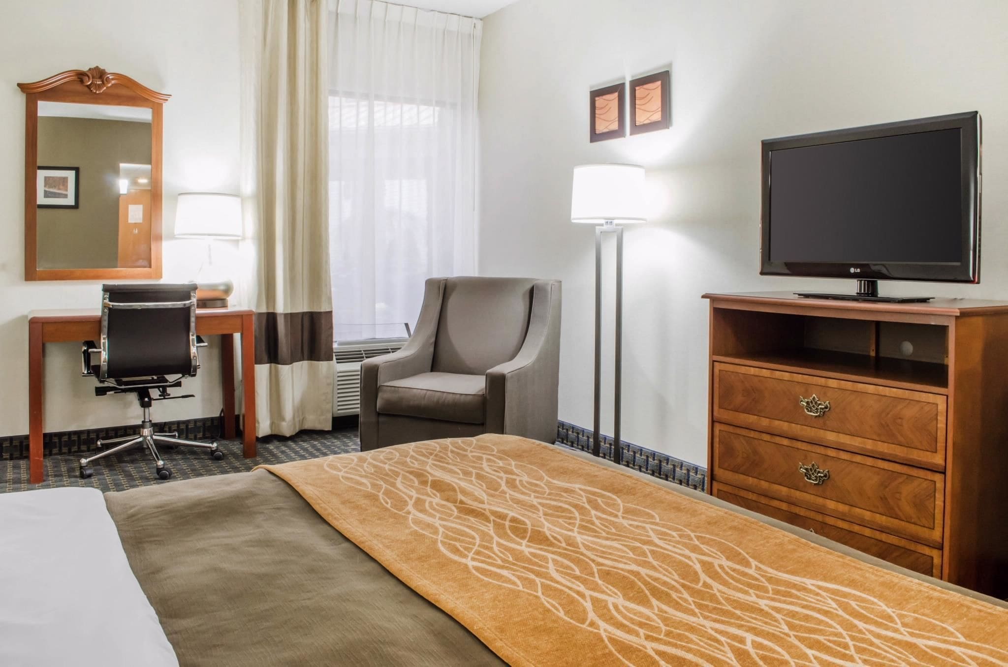 Habitación de hotel con cama, escritorio con silla, televisor sobre un soporte de madera, lámpara y ventana con cortinas.