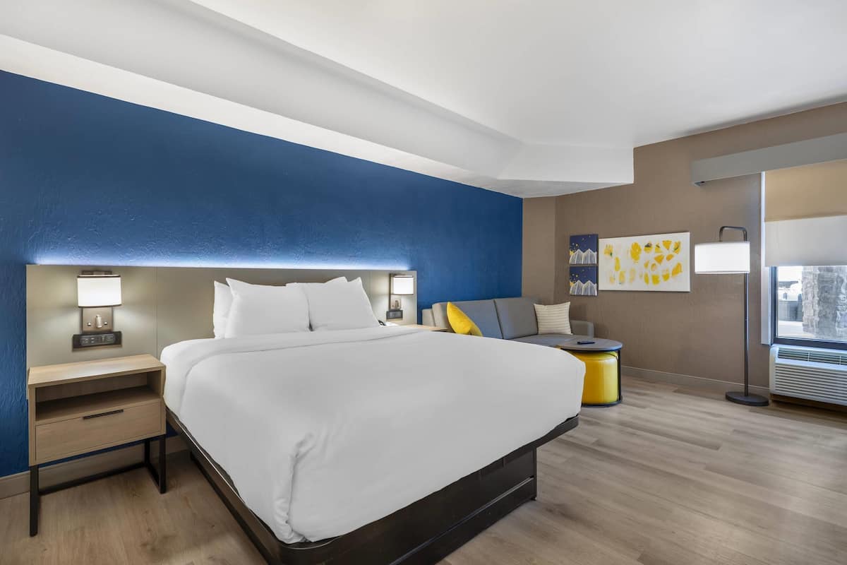 Una habitación de hotel moderna con una cama grande, mesitas de noche, un sofá, una pequeña mesa y obras de arte en las paredes.