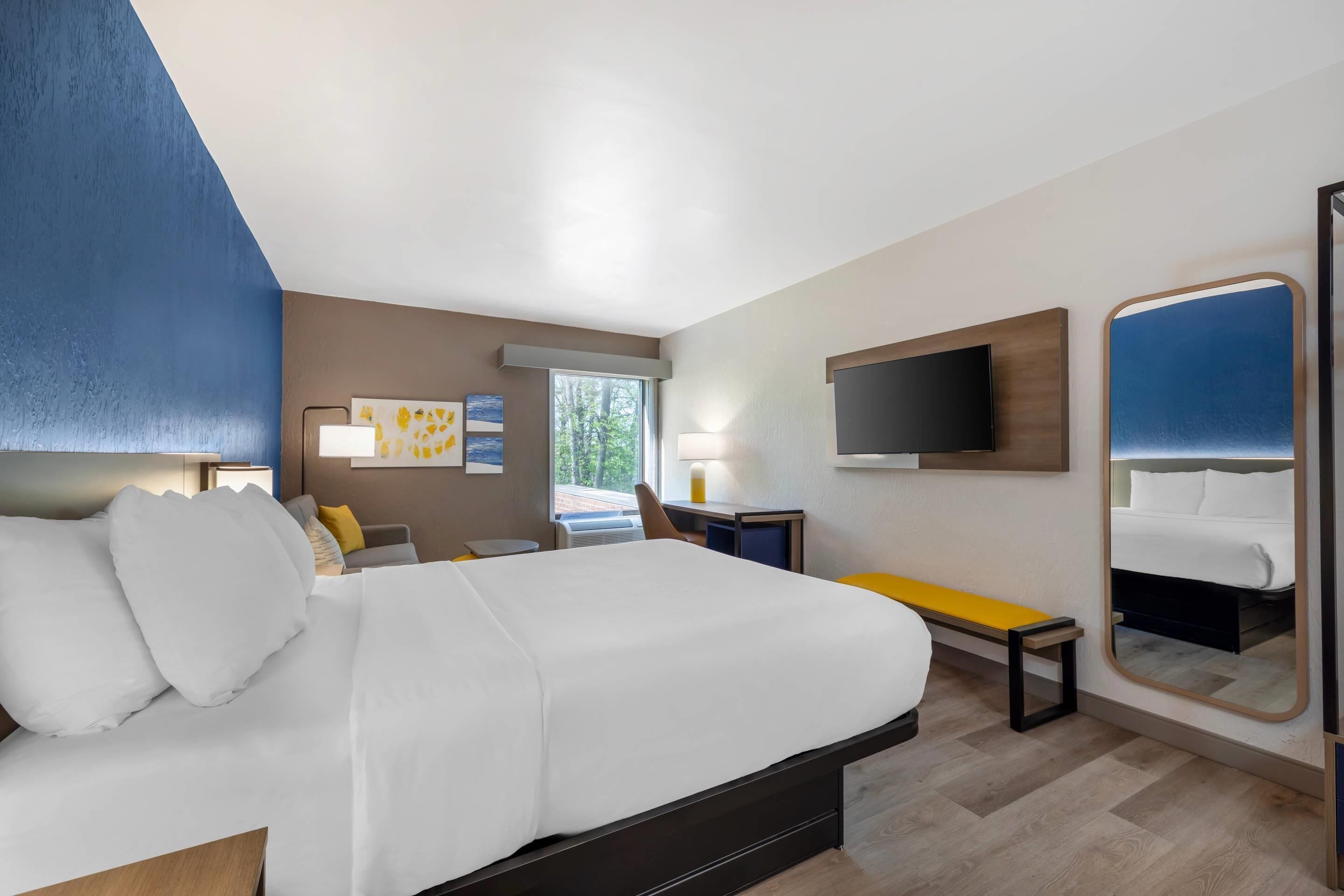 A hotel room with a bed, a desk, a chair, a TV, a mirror, and a window.