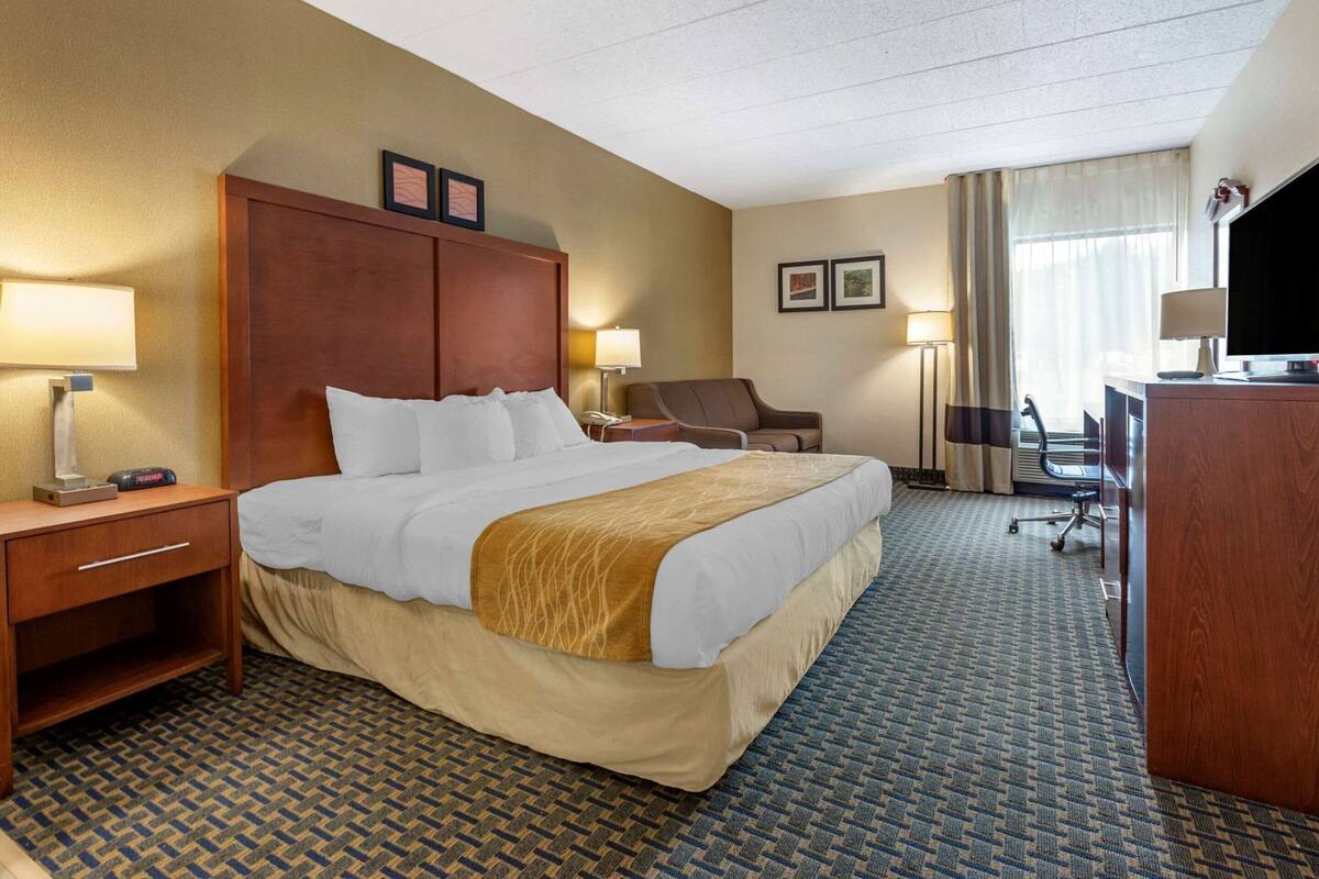 Habitación de hotel con una cama grande, un escritorio, una silla, una lámpara de escritorio, un televisor y un ventanal con cortinas.