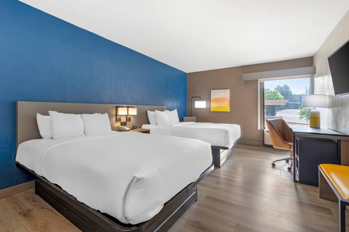 Habitación de hotel con dos camas, un escritorio, una silla, un televisor y un ventanal grande.