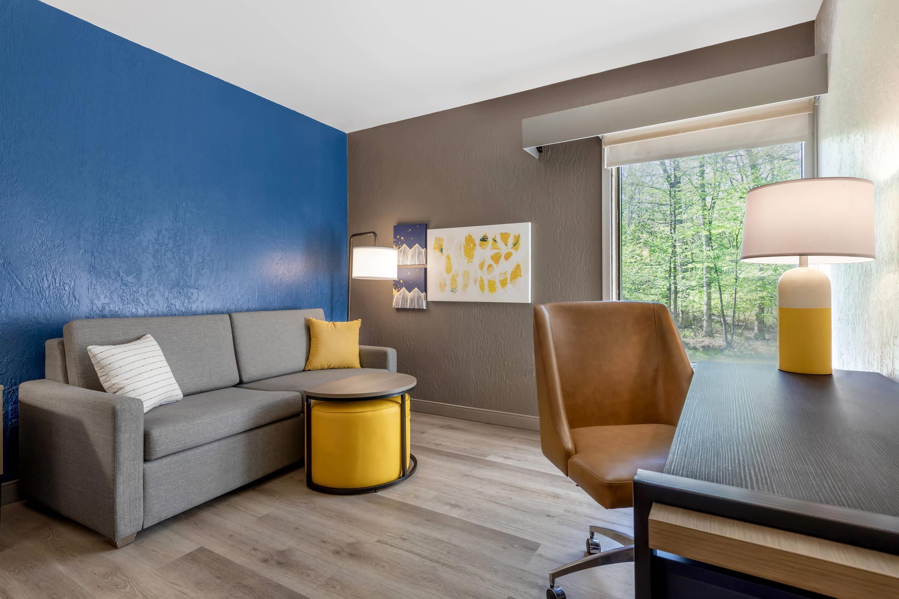 Una habitación de hotel moderna con sofá, una mesa redonda amarilla, una silla marrón, una lámpara y ventana con vista a árboles.
