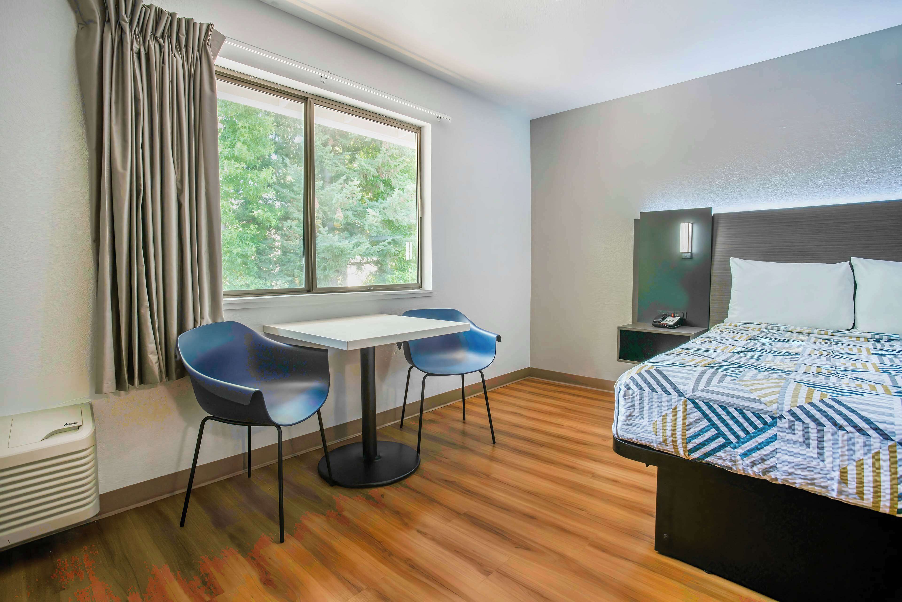 A hotel room with a bed, a small table with two chairs, a window with curtains, and a nightstand with a lamp.