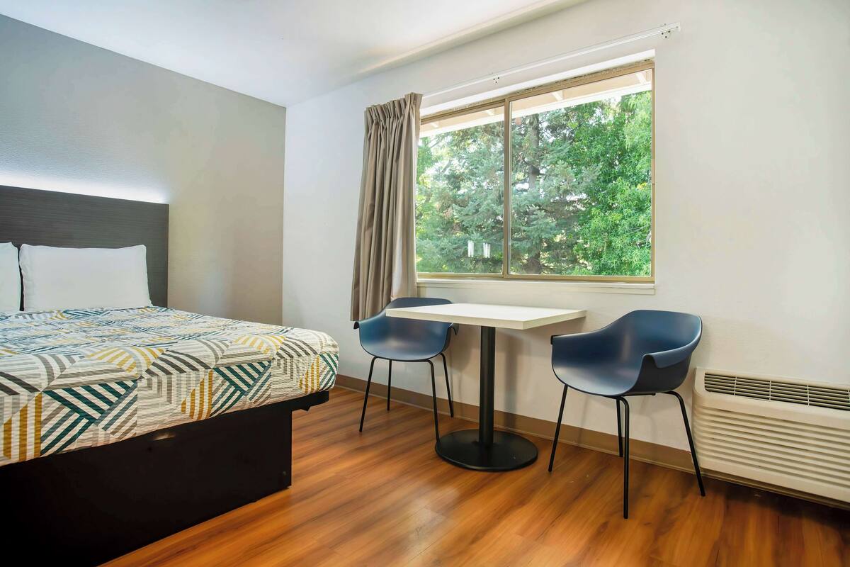 Une chambre d’hôtel avec un lit, une table, deux chaises, une fenêtre avec des rideaux et un radiateur.