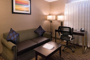 A hotel room with a green sofa, two blue polka dot pillows, a small table with a white folder, a black office chair, a desk with two lamps, and a framed picture on the wall.