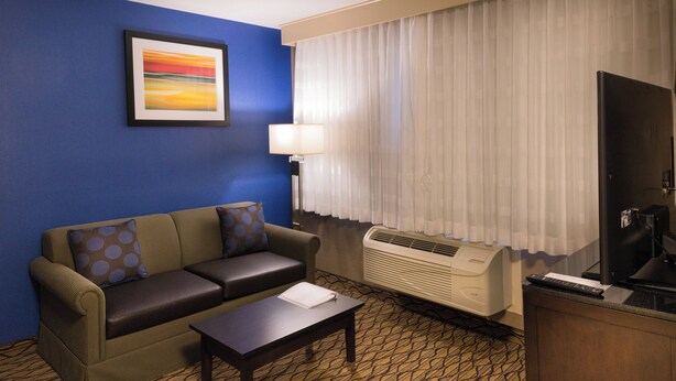 A hotel room with a sofa, a table, a lamp, an air conditioner, and a television. - Holiday Inn Express & Suites Pasadena - Los Angeles by IHG (Pasadena)