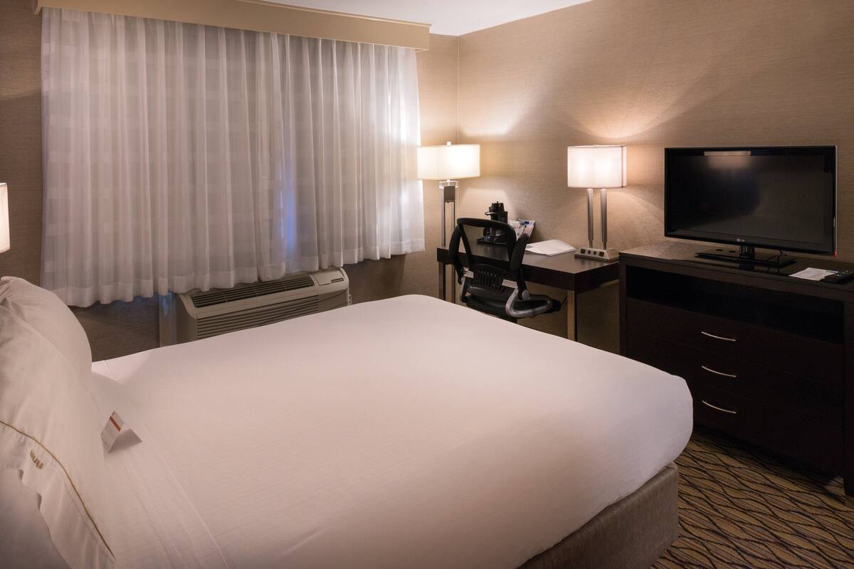 A hotel room with a large bed, a desk with a chair, a flat-screen TV, two lamps, and a window with curtains.