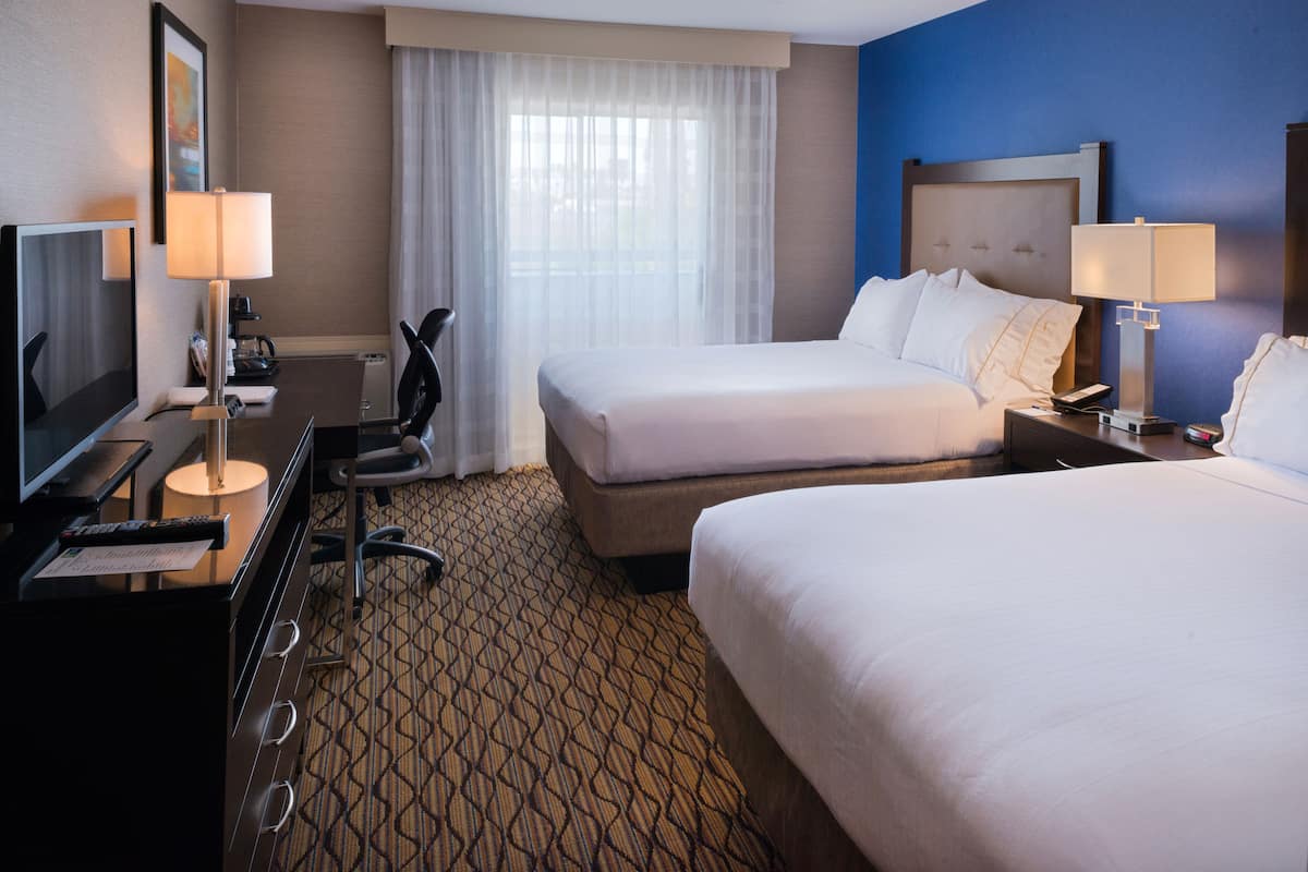 A hotel room with two beds, a desk with a TV, a chair, and a window with sheer curtains.