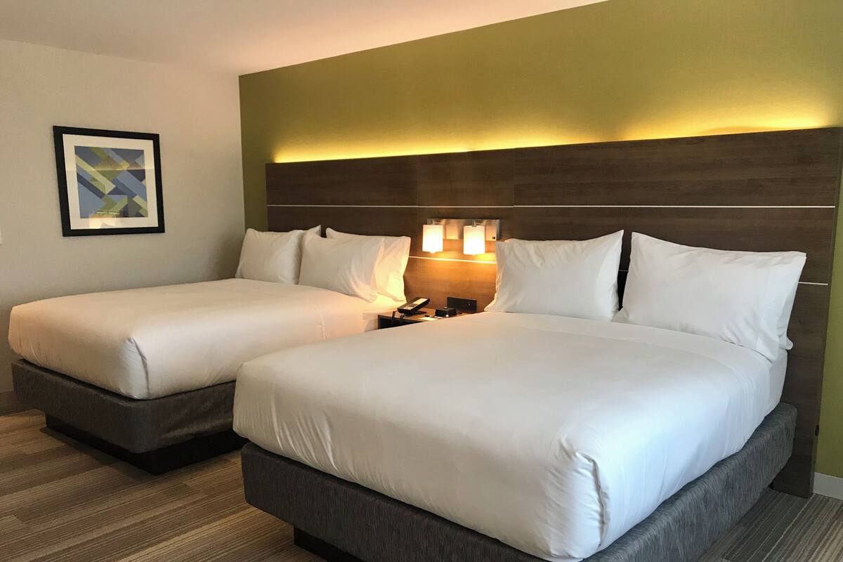 A hotel room with two beds, a headboard with integrated lighting, a bedside table with a lamp, and a framed picture on the wall.