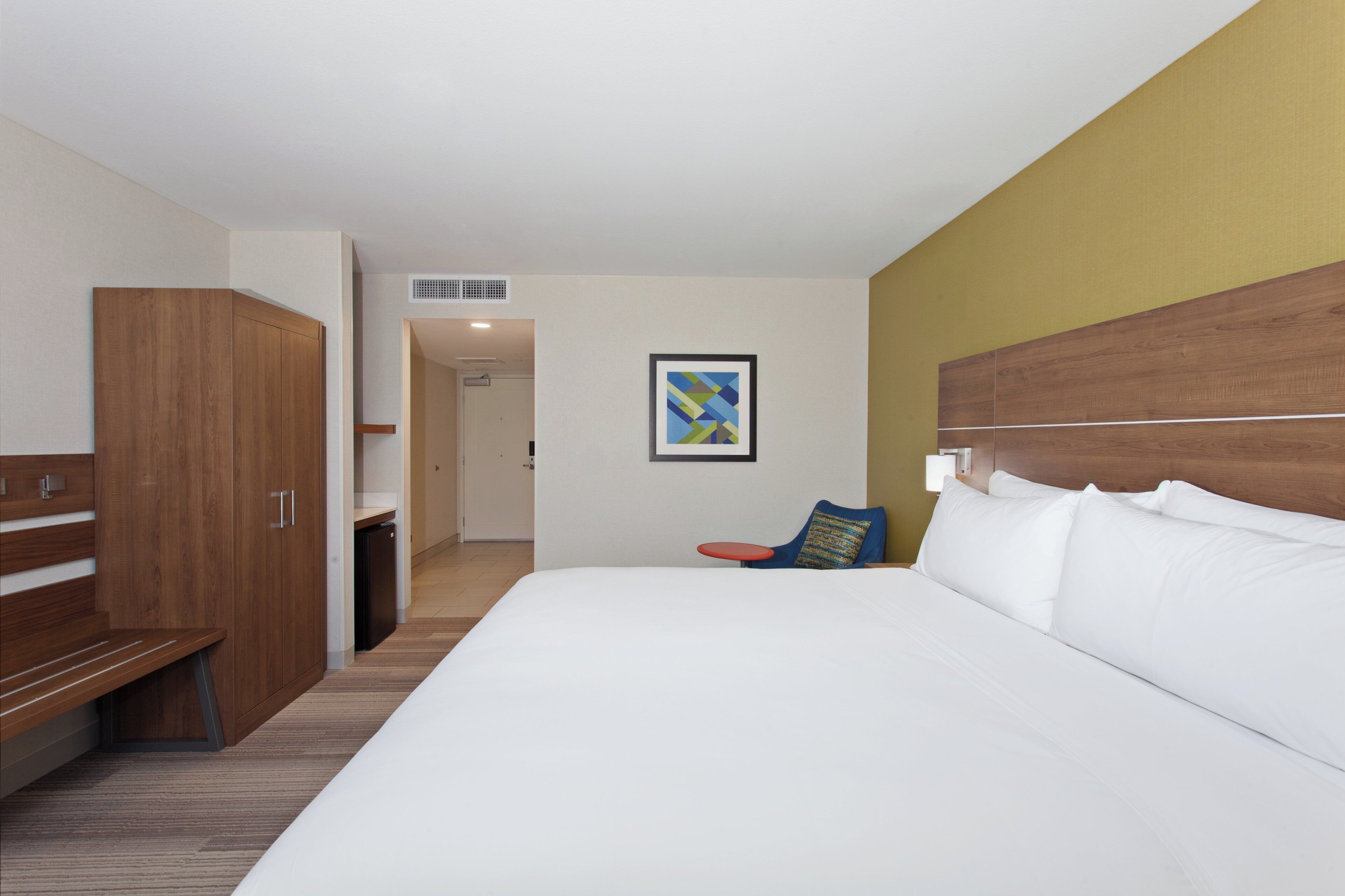 Une chambre d’hôtel avec un grand lit, une armoire en bois, une petite table et une œuvre d’art colorée accrochée au mur.
