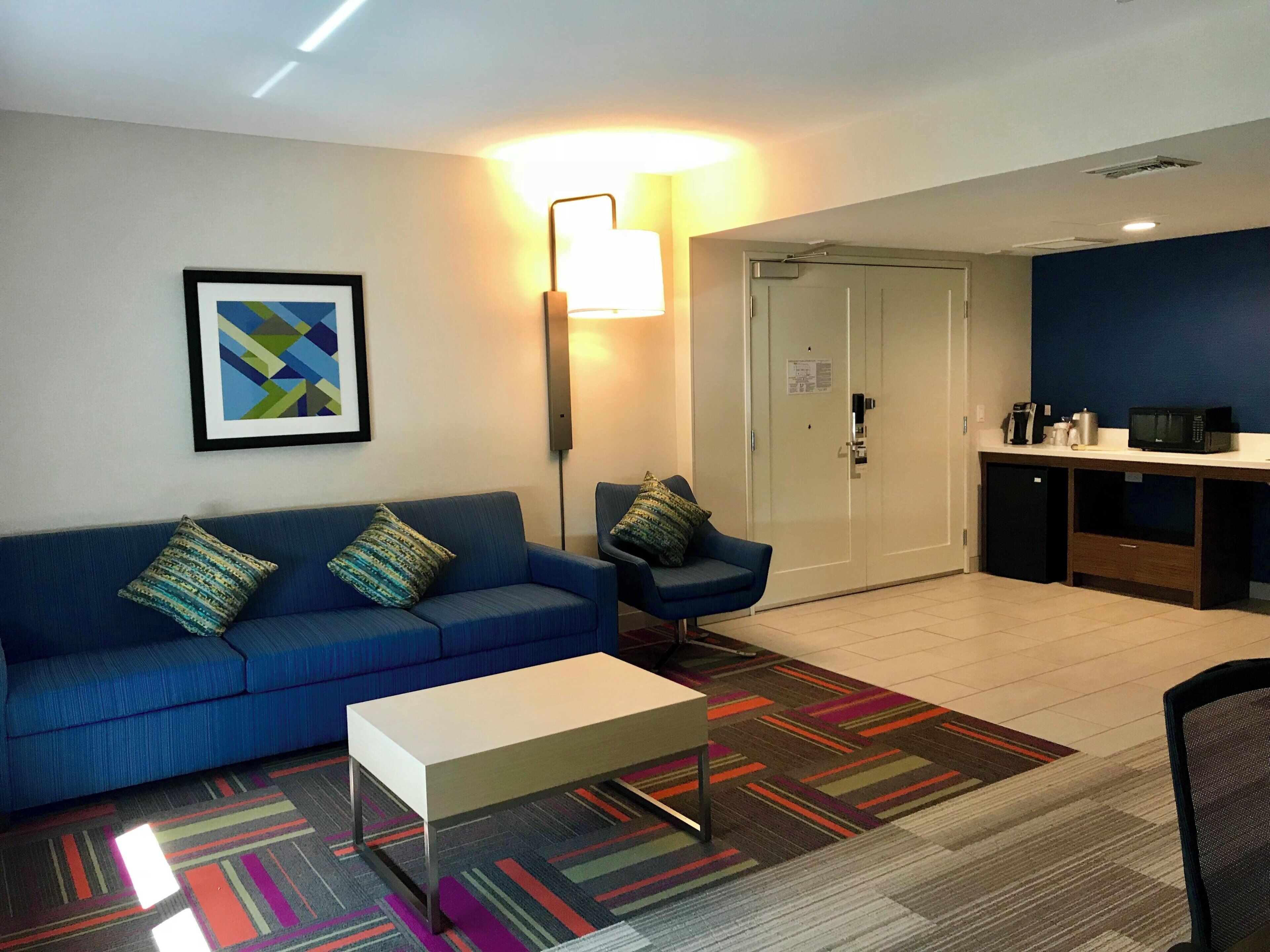 Una habitación de hotel moderna con un sofá azul, una mesa de centro blanca, una alfombra estampada, una obra de arte abstracta enmarcada, un escritorio con microondas y una puerta con cerradura.