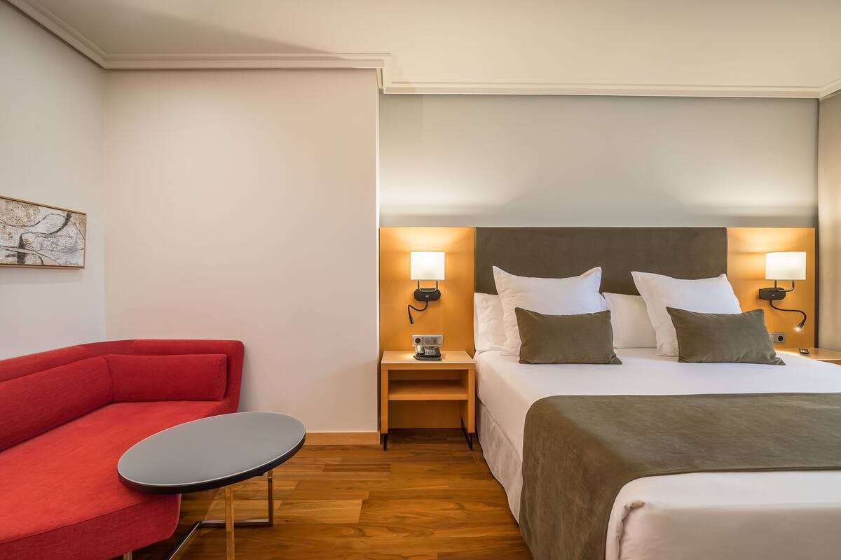 Une chambre d’hôtel avec un lit, un canapé rouge, une petite table et une table de chevet avec une lampe.