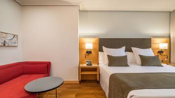 A hotel room with a bed, a red sofa, a small table, and a nightstand with a lamp.