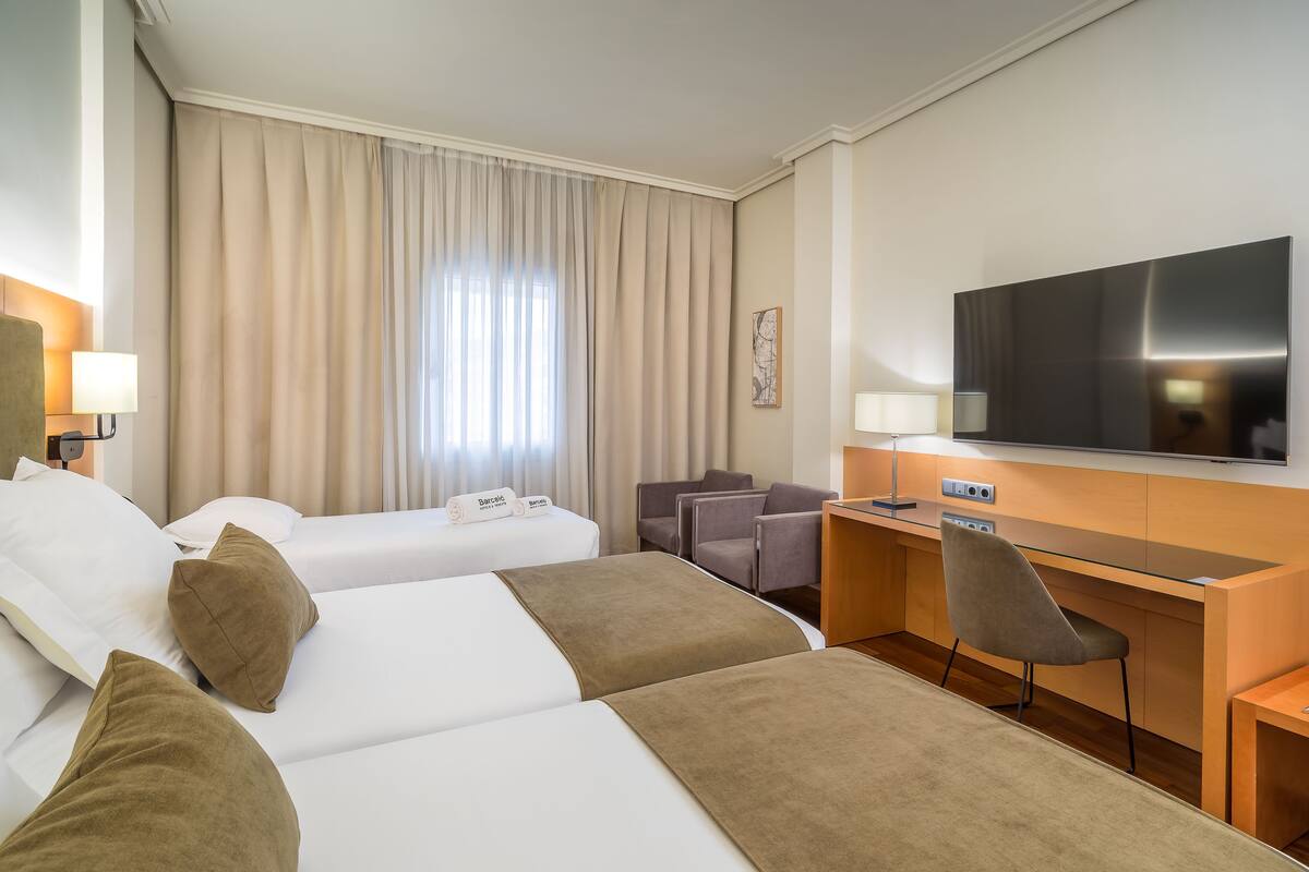 Une chambre d’hôtel avec deux lits, un bureau, une chaise, une télévision et une fenêtre avec des rideaux.