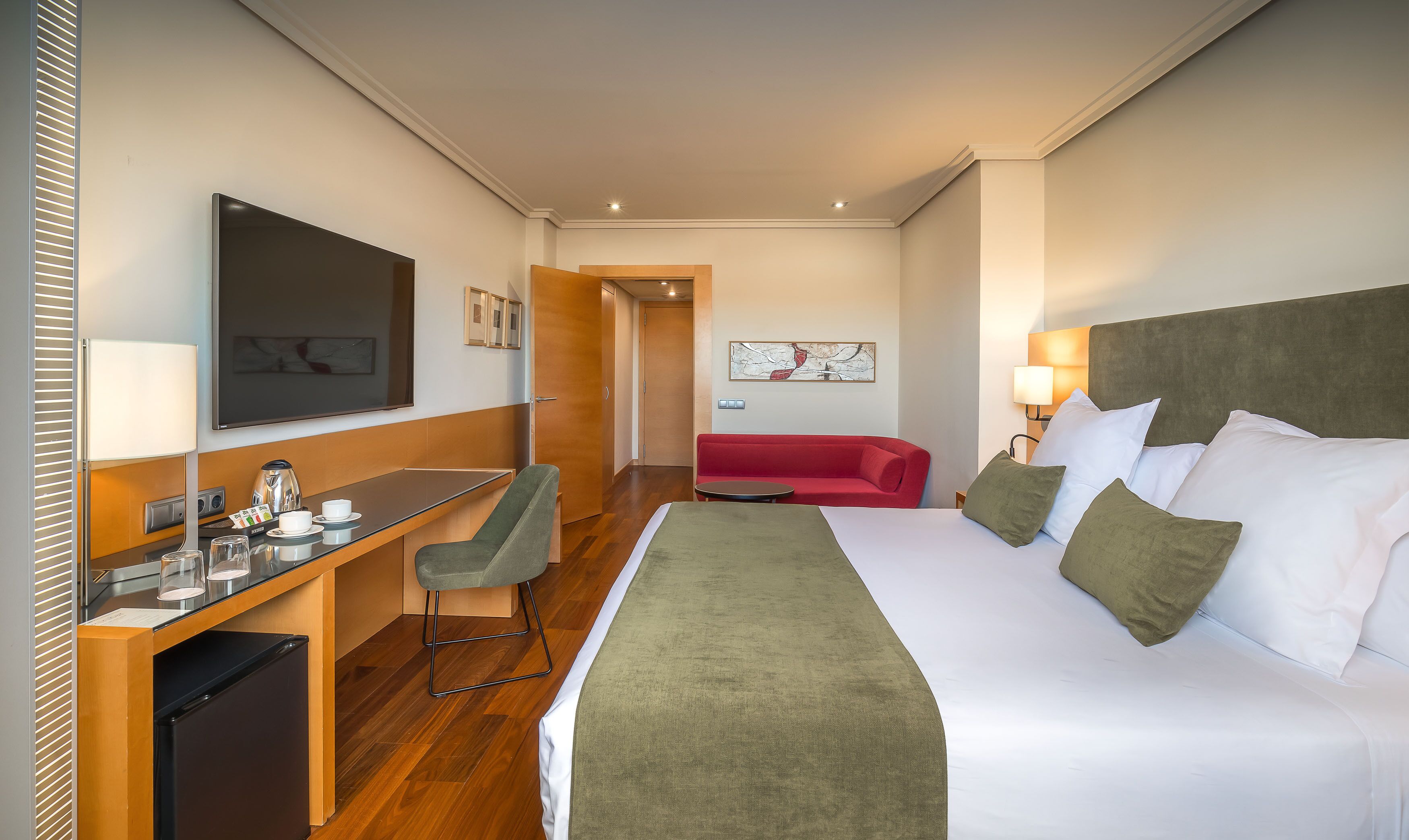 Une chambre d’hôtel avec un lit, un bureau, une chaise, un canapé rouge et un téléviseur.