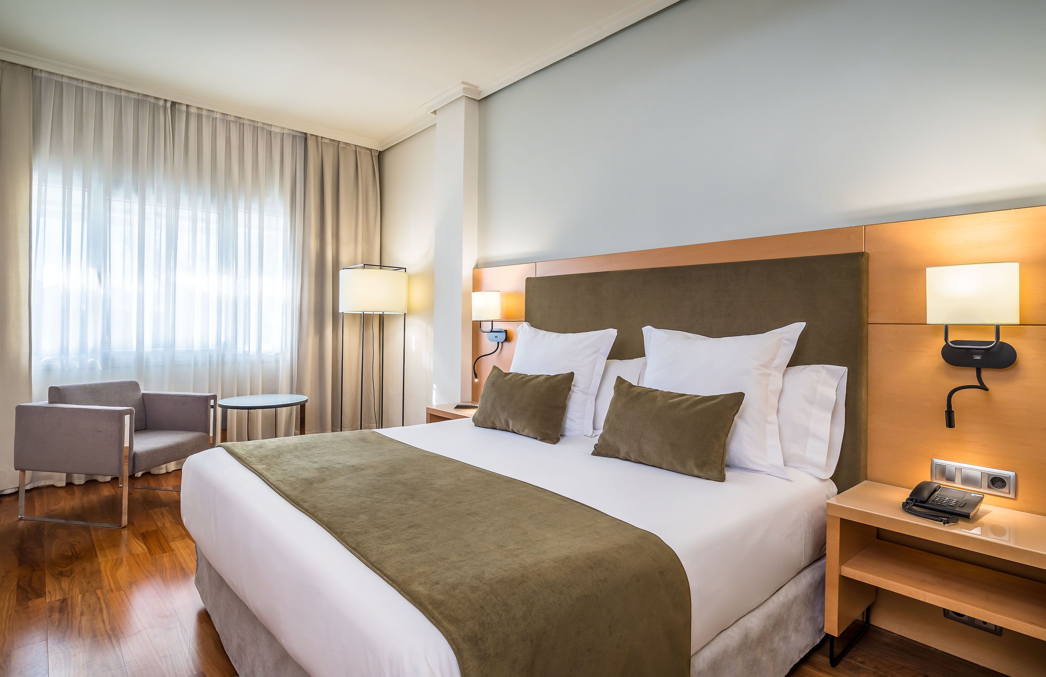 A hotel room with a large bed, two bedside tables with lamps, a chair, and a view of the outdoors through sheer curtains.